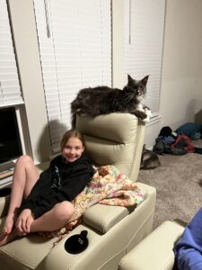 A girl sitting on the arm of a chair with a cat laying on top of it.